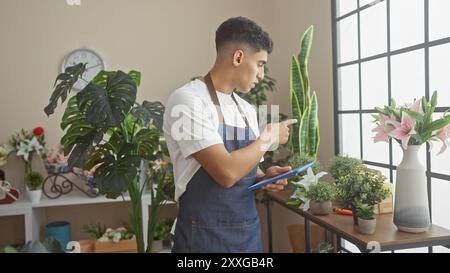 Schöner Mann, der in einem üppigen Blumenladen mit Tablet in der Hand arbeitet, umgeben von lebendiger Flora. Stockfoto