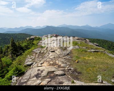 Zwei Wanderer mit roten Rucksäcken begeben sich vom Cascade Mountain hinunter, umgeben von den atemberaubenden Adirondack Mountains unter einem blauen Himmel. Stockfoto