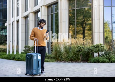 Lächelnder Geschäftsreisender mit Smartphone, während er in urbaner Umgebung den Koffer zieht. Person mit orangefarbenem Pullover, die vor dem Bürogebäude läuft. Erfasst das Konzept von Geschäftsreisen Stockfoto
