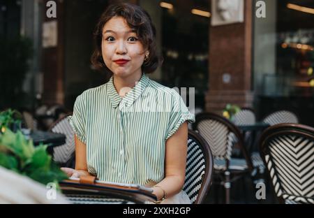 Hübsche asiatische Frau mit kurzen Haaren in grün gestreifter Bluse draußen im Café, hält eine Tablette und macht einen überraschenden Ausdruck Stockfoto