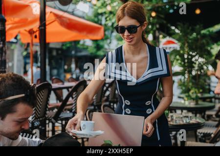 Die Kellnerin serviert einem Gast Kaffee in einem Café im Freien und bietet Gastfreundschaft und Entspannung in einer lebhaften urbanen Umgebung mit orangefarbenen Sonnenschirmen und Stockfoto