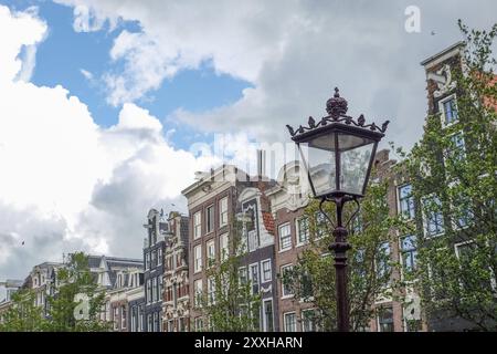 Historische Laterne vor einer Reihe von Gebäuden und Bäumen unter einem geschäftigen Himmel, Amsterdam, Niederlande Stockfoto