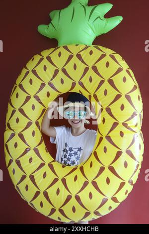 Ein hübsches Kind trägt einen Hut und eine Sonnenbrille, die den Schwimmer auf rotem Hintergrund zum Schwimmen bereit hält. Kopierraum, Tageslicht Stockfoto