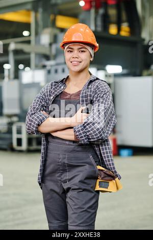 Porträt einer Industriearbeiterin, die selbstbewusst mit gekreuzten Armen in der Fabrik steht, Schutzhelm und Overall trägt und in die Kamera lächelt Stockfoto