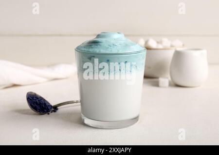 Glas blauer Dalgona Matcha Latte und Teelöffel mit Pudermatcha auf weißem Tisch, Nahaufnahme Stockfoto