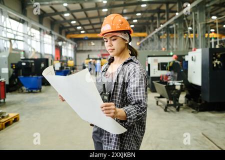 Ingenieur in Schutzhelm, der den Plan in einer geräumigen industriellen Produktionsanlage mit moderner Ausrüstung untersucht Stockfoto