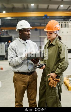 Afroamerikaner diskutieren das Projekt mit einer Ingenieurin, die Sicherheitsausrüstung in einer Industrieanlage trägt und eine Zwischenablage hält und die Aufgabe erklärt Stockfoto