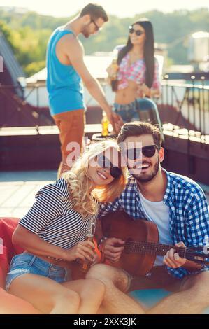 Genießen Sie die Zeit mit Freunden. Wunderschönes junges Paar, das sich aneinander knüpft und mit Gitarre auf dem Sitzsack sitzt, während zwei Leute in der Ba grillen Stockfoto