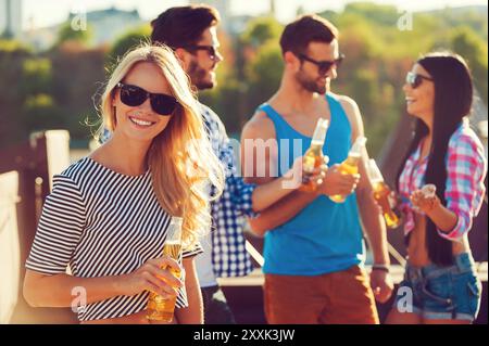 Entspannen Sie sich mit Freunden. Lächelnde junge Frau, die eine Flasche Bier hält und in die Kamera schaut, während drei Leute im Hintergrund miteinander reden Stockfoto