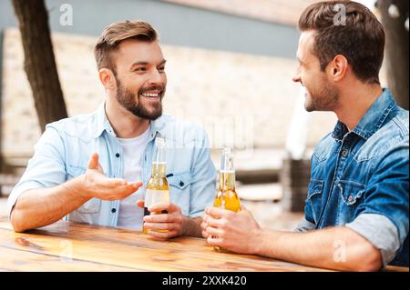Gute Unterhaltung mit einem Freund. Zwei fröhliche junge Männer, die miteinander reden und Flaschen mit Bier halten, während sie draußen stehen Stockfoto