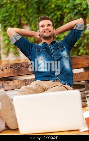 Selbstbewusst und entspannt. Fröhlicher junger Mann, der den Kopf in den Händen hält und lächelt, während er sich draußen an die Holzbank lehnt Stockfoto