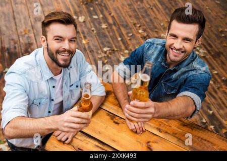 Applaus! Blick von oben auf zwei fröhliche junge Männer, die Flaschen mit Bier ausstrecken und die Kamera betrachten, während sie draußen stehen Stockfoto