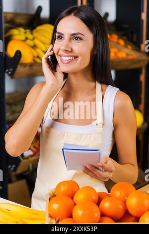 Ordnung bei Früchten. Fröhliche junge Frau in Schürze, die auf dem Handy spricht und Notizblock hält, während sie im Lebensmittelgeschäft mit verschiedenen fru steht Stockfoto