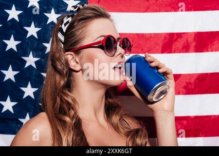 Ein Schluck. Schöne junge Frau, die Sonnenbrille trägt und aus der Dose trinkt, während sie vor der amerikanischen Nationalflagge steht Stockfoto