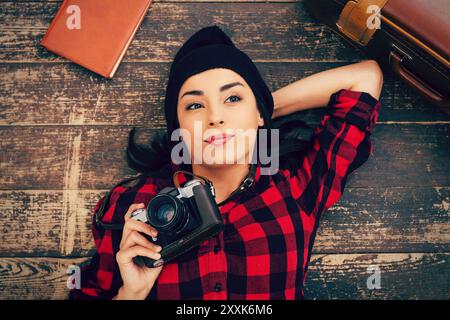 Wunderschöner Tagträumer. Blick von oben auf eine schöne junge Frau in Kopfbedeckung, die auf dem Boden lag und die Kamera hielt, während Koffer und Notizblock in ihrer Nähe lagen Stockfoto