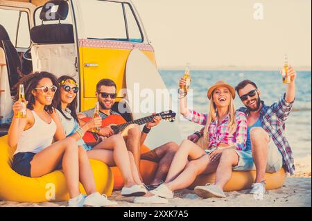 Genießen Sie die gemeinsame Sommerzeit. Gruppe glücklicher junger Leute, die Spaß haben, während sie am Strand in der Nähe ihres Retro-Van sitzen Stockfoto
