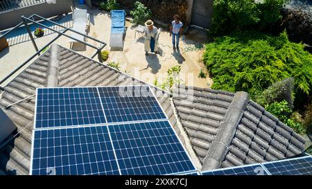 Ein Blick aus der Luft zeigt zwei Frauen in einem sonnigen Hinterhof, eine ist im Garten, während die andere plaudert. Das Dach ist mit Solarpaneelen ausgestattet, Highlight Stockfoto