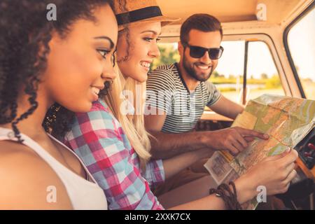 Planung ihrer Roadtrip. Seitenansicht von drei fröhlichen jungen Leuten, die sich die Karte ansehen und lächeln, während sie in ihrem Kleinbus sitzen Stockfoto