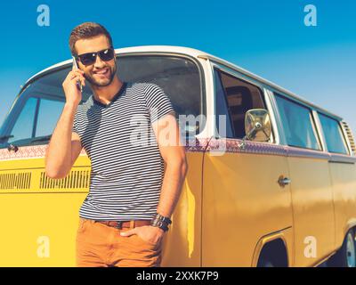 Freunde anrufen. Glücklicher junger Mann, der auf dem Handy spricht und die Hand in der Tasche hält, während er sich an den Retro-Minibus lehnt Stockfoto