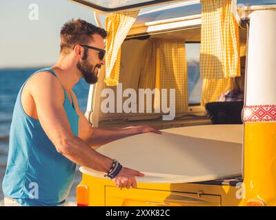 Es wird ein großartiger Tag. Glücklicher junger Mann, der Skimboard aus dem Kofferraum nimmt und am Strand lächelt Stockfoto