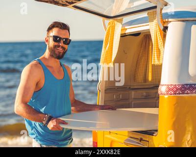 Ich freue mich, am Meer zu sein. Lächelnder junger Mann, der Skimboard aus dem Kofferraum nimmt und die Kamera lächelt, während er am Strand steht Stockfoto