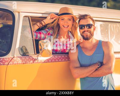 Das Leben ist besser, wenn wir zusammen sind! Fröhliche junge Frau, die in einem Retro-Kleinbus sitzt und durch das Fahrzeugfenster schaut, während der Mann heraussteht Stockfoto