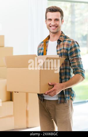 Ich bin froh, in ein neues Haus zu ziehen. Hübscher junger Mann, der eine Pappschachtel hält und in die Kamera lächelt, während andere Kartonschachteln auf dem Hintergrund liegen Stockfoto