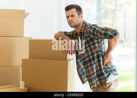 Schmerzen im Rücken. Frustrierter junger Mann, der die Hand auf dem Rücken hält und Negativität ausdrückt, während er sich an den Karton lehnt Stockfoto