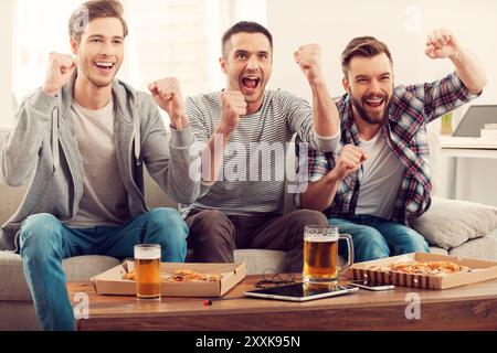 Heimfans. Drei glückliche junge Männer, die Fußballspiele beobachten und die Arme hochhalten, während sie auf dem Sofa sitzen Stockfoto