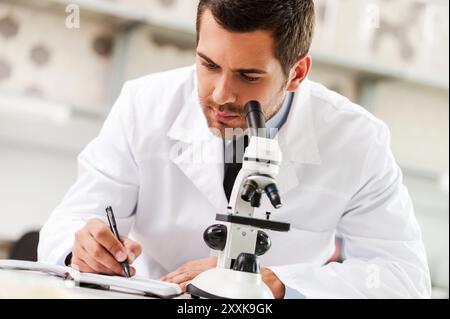 Ich habe mich verpflichtet, das Heilmittel zu finden. Gutaussehender junger Wissenschaftler in weißer Uniform mit Mikroskop und Notizblock, während er am Arbeitsplatz sitzt Stockfoto