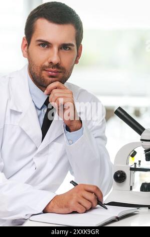 Selbstbewusster Wissenschaftler. Hübscher junger Wissenschaftler in weißer Uniform, der Hand am Kinn hält und in Notizblock schreibt, während er an seinem Arbeitsplatz sitzt Stockfoto