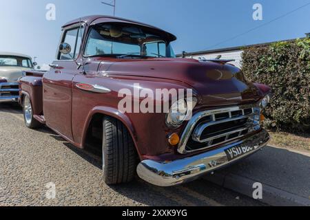 Rayleigh, Großbritannien. August 2024. Ein kastanienbrauner Pickup-Truck aus der Mitte des 20. Jahrhunderts mit Chrom-Stoßfänger und Kühlergrill, teilweise auf einem Bordstein geparkt. Das Fahrzeug verfügt über runde Scheinwerfer und ein unverwechselbares Vintage-Design. Ein weiterer Oldtimer ist im Hintergrund zu sehen, mit einer Hecke und einem Gebäude, das die Vorstadtszene abrundet. Amerikanisches Auto und Truck treffen sich in der Farm Fresh Garage. Bei der Veranstaltung können die Besitzer ihre Fahrzeuge mitbringen, damit die Besucher einen Blick aus der Nähe werfen können. Penelope Barritt/Alamy Live News Stockfoto