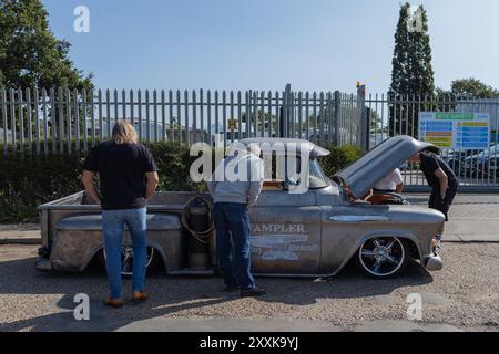 Rayleigh, Großbritannien. August 2024. Ein verwitterter Vintage-Pickup mit kundenspezifischen Rädern und Metallic-Finish wird von drei Personen während einer britischen Automobilveranstaltung genau geprüft. Der Lkw verfügt über ein sichtbares Branding („TRAMPLER“) und eine offene Motorhaube, was auf mechanische Interessen oder eine Anzeige hinweist. Amerikanisches Auto und Truck treffen sich in der Farm Fresh Garage. Bei der Veranstaltung können die Besitzer ihre Fahrzeuge mitbringen, damit die Besucher einen Blick aus der Nähe werfen können. Penelope Barritt/Alamy Live News Stockfoto