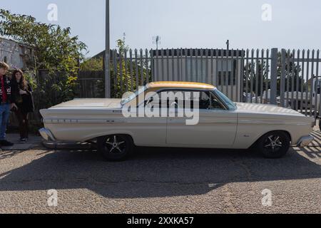 Rayleigh, Großbritannien. August 2024. Ein klassisches weißes Mercury-Auto mit gelbem Dach und schwarzen Rädern mit Chromakzenten, parkt neben einem Metallzaun in einem Industriegebiet. Das Design des Fahrzeugs erinnert an ein Modell aus den 1960er Jahren, und mehrere Personen sind am Heck sichtbar. Die Szene erinnert an Americana aus der Mitte des Jahrhunderts und an Nostalgie in der Automobilbranche. Amerikanisches Auto und Truck treffen sich in der Farm Fresh Garage. Bei der Veranstaltung können die Besitzer ihre Fahrzeuge mitbringen, damit die Besucher einen Blick aus der Nähe werfen können. Penelope Barritt/Alamy Live News Stockfoto