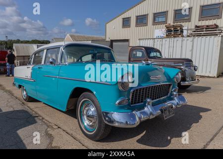 Rayleigh, Großbritannien. August 2024. Ein türkisfarbener und weißer Chevrolet Bel Air aus den 1950er Jahren parkte vor einem beigefarbenen Industriegebäude, begleitet von einem dunklen braunen Fahrzeug aus den 1940er Jahren. Die Szene zeigt eine Zusammenkunft von Oldtimer-Fans und zeigt die amerikanische Automobil-Design- und Restaurierungskultur der Mitte des Jahrhunderts in einem britischen Umfeld. Amerikanisches Auto und Truck treffen sich in der Farm Fresh Garage. Bei der Veranstaltung können die Besitzer ihre Fahrzeuge mitbringen, damit die Besucher einen Blick aus der Nähe werfen können. Penelope Barritt/Alamy Live News Stockfoto