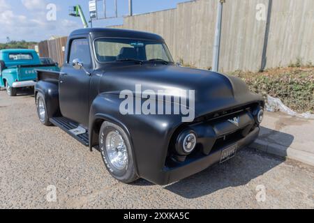 Rayleigh, Großbritannien. August 2024. Ein mattschwarzer Pickup-Truck aus der Mitte des 20. Jahrhunderts mit Chromrädern und einer maßgeschneiderten Karosserie steht neben einer Betonmauer unter klarem Himmel. Im Hintergrund ist ein türkisfarbener Oldtimer zu sehen. Die Szene erinnert an die klassische Automobil-Design- und Restaurierungskultur, die in einem britischen Stadtumfeld festgehalten wurde. Amerikanisches Auto und Truck treffen sich in der Farm Fresh Garage. Bei der Veranstaltung können die Besitzer ihre Fahrzeuge mitbringen, damit die Besucher einen Blick aus der Nähe werfen können. Penelope Barritt/Alamy Live News Stockfoto