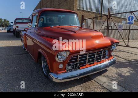 Rayleigh, Großbritannien. August 2024. Ein klassischer roter Pickup mit Chromdetails steht auf einer gepflasterten Fläche in der Nähe eines umzäunten Industriegebiets. Ein zweiter Oldtimer-Lkw ist dahinter sichtbar. Die Szene erfasst das Automobil-Design der Mitte des 20. Jahrhunderts in einem modernen industriellen Kontext und hebt die Kultur der Restaurierung und den urbanen Kontrast hervor. Amerikanisches Auto und Truck treffen sich in der Farm Fresh Garage. Bei der Veranstaltung können die Besitzer ihre Fahrzeuge mitbringen, damit die Besucher einen Blick aus der Nähe werfen können. Penelope Barritt/Alamy Live News Stockfoto