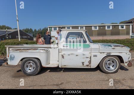 Rayleigh, Großbritannien. August 2024. Ein weißer Pickup-Lkw aus der Mitte des 20. Jahrhunderts mit sichtbarem Rost und Lackverschleiß steht auf einer gepflasterten Fläche in der Nähe eines Industriebaus aus Wellblech. Die alte Karosserie des Lkws steht im Kontrast zu den glänzenden modernen Rädern. Vier Personen stehen in der Nähe unter einem klaren blauen Himmel, was auf ein zwangloses Treffen oder eine Gemeindeversammlung hindeutet. Amerikanisches Auto und Truck treffen sich in der Farm Fresh Garage. Bei der Veranstaltung können die Besitzer ihre Fahrzeuge mitbringen, damit die Besucher einen Blick aus der Nähe werfen können. Penelope Barritt/Alamy Live News Stockfoto