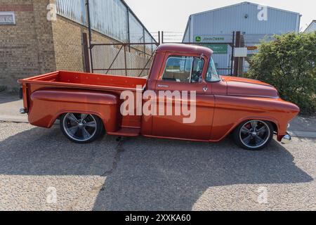 Rayleigh, Großbritannien. August 2024. Ein Chevrolet Pickup-Truck aus der Mitte des 20. Jahrhunderts mit einer speziell angefertigten orangefarbenen Lackierung, abgesenkter Federung und modernen Leichtmetallrädern, der in der Nähe eines Industrielagers mit „John Deere“-Schildern parkt. Die Szene stellt klassisches Automobil-Design mit moderner Hot Rod-Kultur in einem britischen urbanen Setting gegenüber. Amerikanisches Auto und Truck treffen sich in der Farm Fresh Garage. Bei der Veranstaltung können die Besitzer ihre Fahrzeuge mitbringen, damit die Besucher einen Blick aus der Nähe werfen können. Penelope Barritt/Alamy Live News Stockfoto