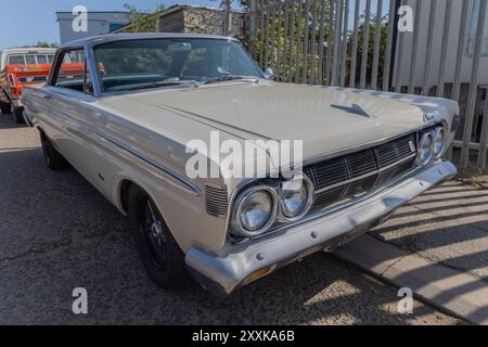 Rayleigh, Großbritannien. August 2024. Ein gut erhaltener weißer Mercury Comet, der in der Nähe eines Metallzaun parkt und das amerikanische Automobildesign der Mitte des 20. Jahrhunderts zeigt. Das Fahrzeug verfügt über Chromdetails, zwei runde Scheinwerfer und eine polierte Karosserie. Ein zweiter Oldtimer, möglicherweise ein orangener Kombi, ist teilweise im Hintergrund zu sehen. Amerikanisches Auto und Truck treffen sich in der Farm Fresh Garage. Bei der Veranstaltung können die Besitzer ihre Fahrzeuge mitbringen, damit die Besucher einen Blick aus der Nähe werfen können. Penelope Barritt/Alamy Live News Stockfoto