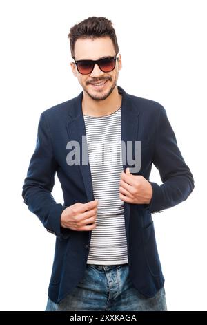 Elegant und stilvoll. Hübscher junger Mann mit Sonnenbrille, der seine Jacke anpasst, während er vor weißem Hintergrund steht Stockfoto