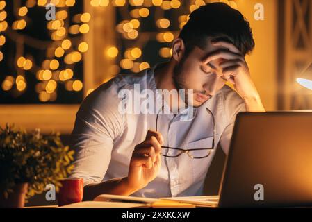 Erschöpft. Frustrierter junger Mann, der eine Brille trägt und die Augen geschlossen hält, während er nachts mit Weihnachtslilie an seinem Arbeitsplatz sitzt Stockfoto