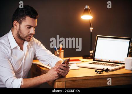 Immer in Kontakt. Selbstbewusster junger Mann, der nachts an seinem Arbeitsplatz sitzt Stockfoto