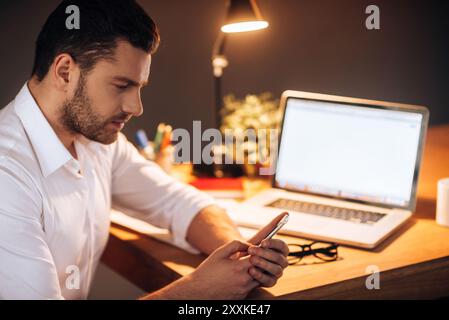 Bleiben Sie auch spät in Kontakt. Selbstbewusster junger Mann, der nachts auf sein Smartphone schaut, während er am Arbeitsplatz sitzt Stockfoto