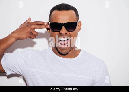 Knall! Wütender junger afrikanischer Mann in Sonnenbrille, der Fingerpistole nahe dem Kopf und Mund offen hält, während er vor weißem Hintergrund steht Stockfoto