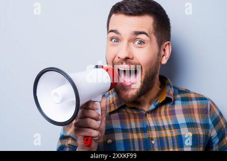 Sie möchten gehört werden. Hübscher junger Mann, der durch ein Megaphon schreit, während er vor grauem Hintergrund steht. Stockfoto