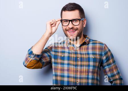 Gut aussehend in Brillen. Hübscher junger Mann, der die Brille anpasst und lächelt, während er vor grauem Hintergrund steht Stockfoto