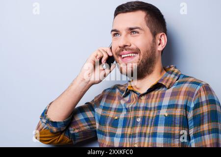 Sprechen Sie gerne. Hübscher junger Mann im Hemd, der das Handy hält und wegblickt, während er vor grauem Hintergrund steht Stockfoto