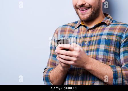Die richtigen Wörter auswählen. Nahaufnahme eines gutaussehenden jungen Mannes im Hemd, der Handy hält und lächelt, während er vor grauem Hintergrund steht Stockfoto