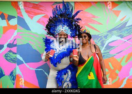 EDITORIAL USE ONLY Carnival-Besucher Michael Styles und Sereka Robert vor einem Kunstwerk des lokalen Straßenkünstlers CEE im Nisa-Shop in Ladbroke Grove, London. Bilddatum: Sonntag, 25. August 2024. Hennessy hat sich mit einer Reihe unabhängiger Eckläden entlang der Paraderoute zusammengetan, um den Notting Hill Carnival und die Markteinführung der limitierten Flasche zu feiern. Das Foto sollte lauten: Matt Crossick/PA Media Assignments Stockfoto